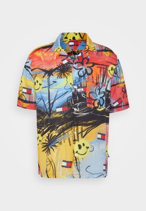 Homme portant une chemise florale colorée, un pantalon noir avec des imprimés smiley, et des baskets blanches à motifs, debout dans une pièce avec des tabourets en bois et des étagères de bouteilles.