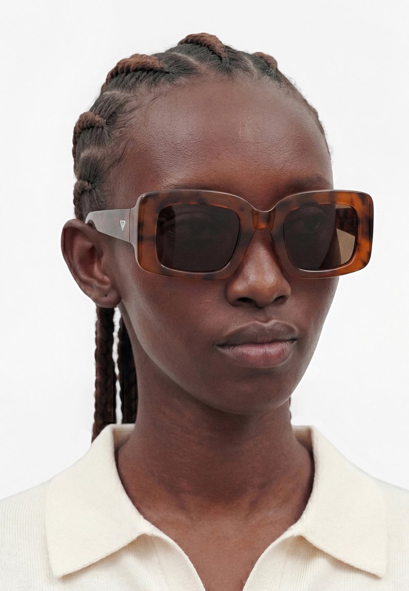 Femme aux cheveux tressés portant de grandes lunettes de soleil rectangulaires écaille de tortue et une chemise claire à col.