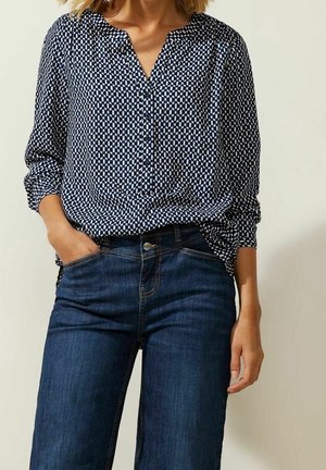 Kobieta ubrana w bluzkę z długim rękawem w granatowo-biały geometryczny wzór, wpuszczoną w ciemnoniebieskie dżinsy, z jedną ręką w kieszeni.
