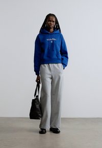 Modrá mikina s kapucňou so sťahovacími šnúrkami a logom, doplnená sivými teplákmi. Čierna kabelka a topánky dotvárajú vzhľad. Hladké textúry, voľný strih.