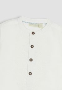 Witte katoenen shirt met een ronde hals, centrale knopen sluiting en bloemengebreide details. Heeft vijf decoratieve bruine knopen.