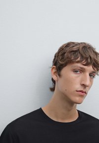 Černé bavlněné tričko s kulatým výstřihem, na němž je mladá osoba s vlnitými hnědými vlasy, na světlém šedém pozadí.