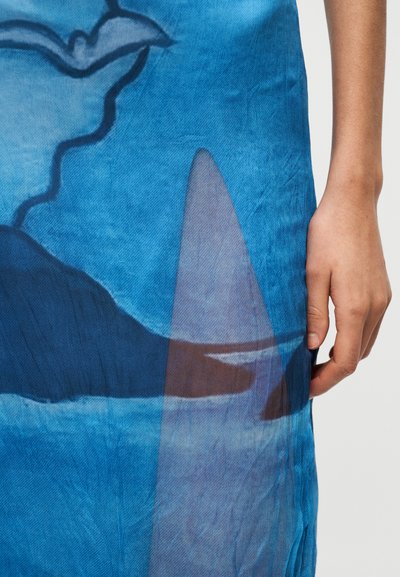 Το ύφασμα με μπλε σχέδια παρουσιάζει αφηρημένα μοτίβα με διαφορετικές αποχρώσεις του μπλε και λεπτές υφές. Ένα χέρι αγγίζει την άκρη, επιδεικνύοντας το υλικό.