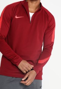 Haut Nike rouge à manches longues avec une ouverture zippée au quart, présentant des accents roses sur les épaules et une texture de tissu lisse et extensible.