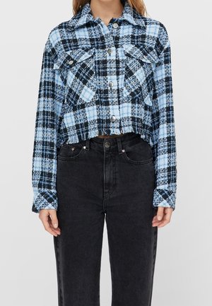 Vrouw draagt een cropped blauw-zwart geruit blouse met een rafelige zoom en een zwarte high-waist jeans.