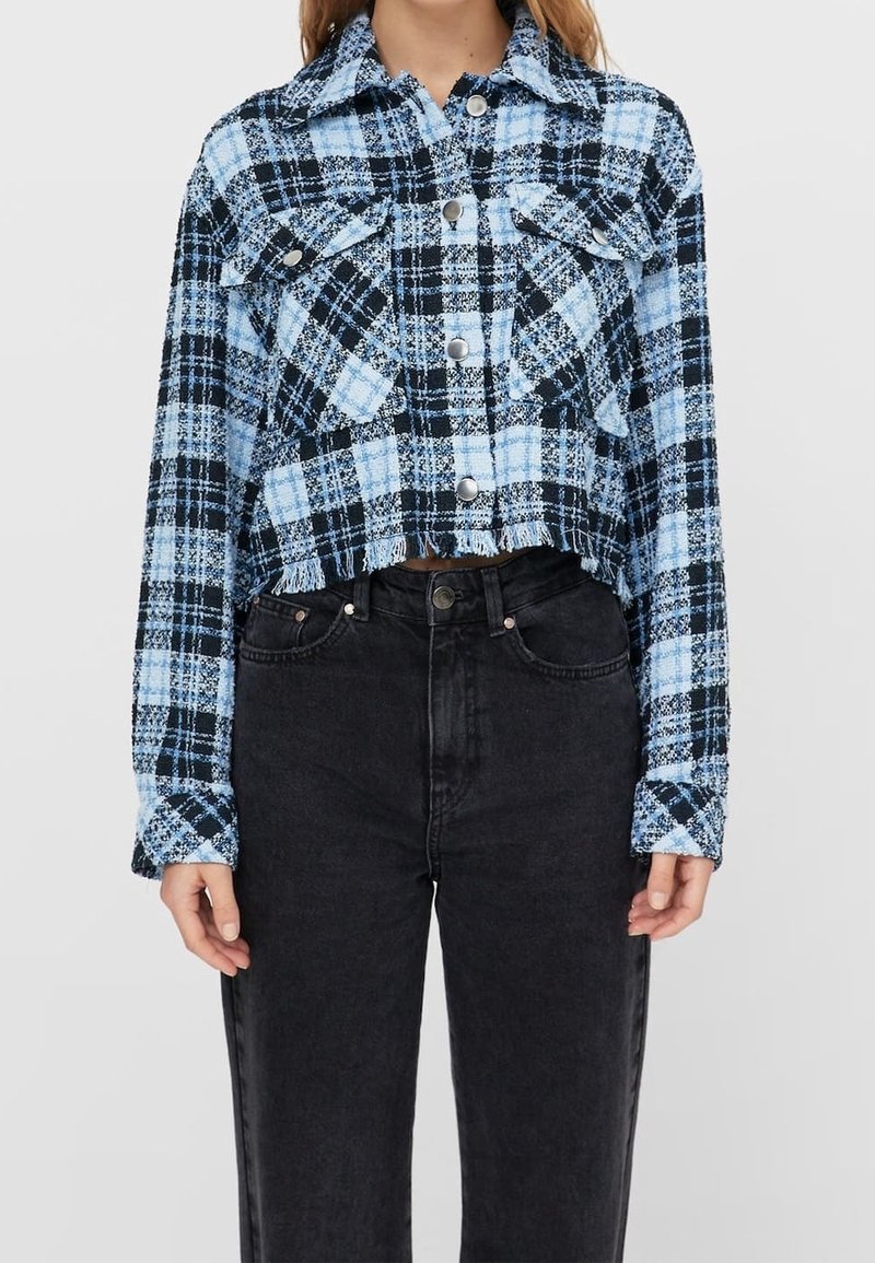 Femme portant une chemise courte à carreaux bleus et noirs avec ourlet effiloché et jean noir taille haute.