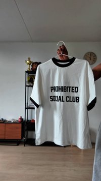 Balta medvilninė marškinėliai su juodu trikotažu apykaklės ir rankovių krašteliuose, ant kurių užrašyta "PROHIBITED SOCIAL CLUB" ryškiu juodu šriftu.