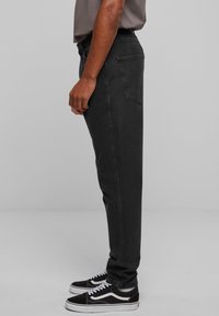 Asmuo stovintis šonu, dėvintis juodus siaurus džinsus, pilką marškinėlį ir juodai baltus sportbačius prieš paprastą pilką foną.