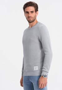 Hellgrauer Strickpullover mit Rundhalsausschnitt, gerippten Bündchen und Saum. Verfügt über ein strukturiertes Muster und ein Etikett in der unteren linken Ecke.