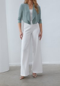 Světle modrý zkrácený cardigan přes bílý top s úzkými ramínky a široké bílé kalhoty. Oblečení doplňují stříbrné akcenty na vysokých podpatkách.