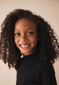 Niño sonriente con cabello rizado, vestido con un suéter de cuello alto negro acanalado. Fondo claro que resalta los rasgos faciales y la textura del cabello.