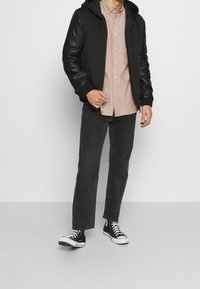 Zwarte hoodie met leren mouwen, beige overhemd met knopen, donkergrijze jeans en zwarte high-top sneakers. Eenvoudige, casual outfit.