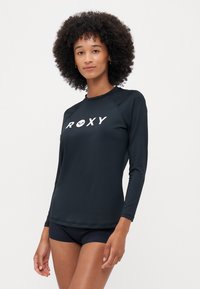 Melna ilga piedurkne rash guard, kas izgatavots no elastīga materiāla, ar baltu "ROXY" logotipu. Viegls, piegulošs dizains ar plakano šuvēm.