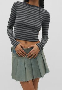 Jeune personne aux cheveux tressés portant un haut gris rayé à manches longues raccourci et une mini-jupe plissée en denim bleu clair.