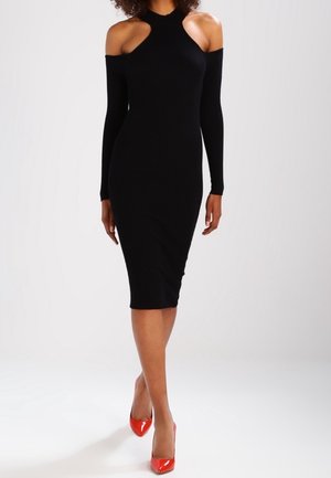 Schwarzes geripptes Strickkleid mit schulterfreiem Design, langen Ärmeln und taillierter Silhouette. Kombiniert mit glänzenden roten High Heels.