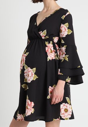 Schwangere Person trägt ein schwarzes Blumenkleid mit pinkfarbenen Blumen, mit V-Ausschnitt und mehrlagigen Glockenärmeln.