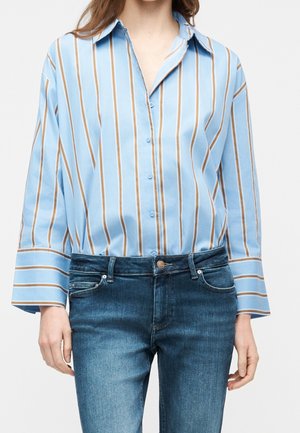Femme portant une chemise bleu clair à manches longues avec des rayures verticales marron et blanches et un jean en denim bleu.