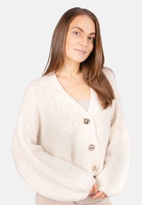 Elfenbeinfarbener, gestrickter Cardigan mit V-Ausschnitt, übergroßen Ärmeln und drei dekorativen Knöpfen vorne, der ein strukturiertes, grobmaschiges Muster zeigt.