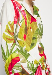Bluse mit Blumenmuster, die lebhafte rote, grüne und gelbe Blätter zeigt, aus leichtem Stoff mit einem geknoteten Detail an der Taille.