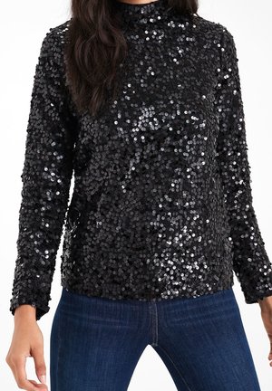 Haut noir à manches longues recouvert de sequins brillants porté avec un jean bleu foncé, montrant le torse et les bras sur fond blanc.