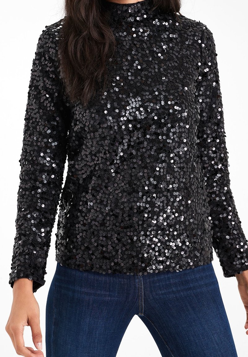 Haut noir à manches longues recouvert de sequins brillants porté avec un jean bleu foncé, montrant le torse et les bras sur fond blanc.