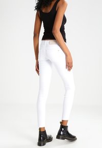 Jean skinny blanc avec détail de poche arrière, associé à des bottines vernies noires et un débardeur noir, sur un fond uni.