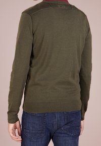 Man med olivgrön långärmad tröja och mörkblå jeans, sedd bakifrån mot en beige bakgrund.