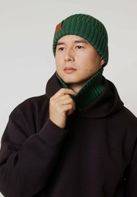 Zielona, prążkowana dzianinowa czapka z tagiem ze skóry, w zestawie z dopasowanym kominem oraz ciemną bluzą z kapturem, prezentująca miękką teksturę i przylegające dopasowanie.