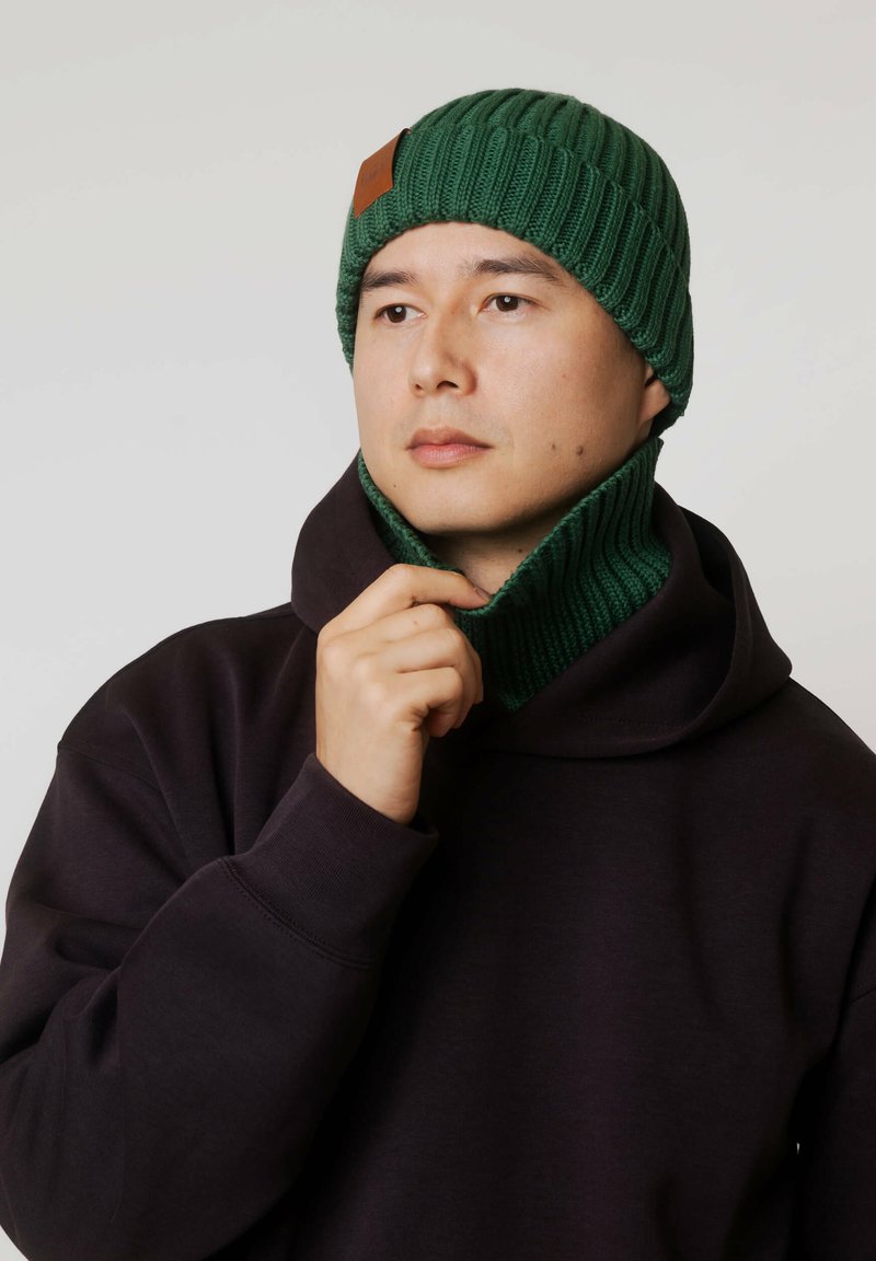 Zielona, prążkowana dzianinowa czapka z tagiem ze skóry, w zestawie z dopasowanym kominem oraz ciemną bluzą z kapturem, prezentująca miękką teksturę i przylegające dopasowanie.