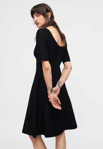 Άτομο με τατουάζ που φοράει μαύρο φόρεμα με κοντά μανίκια, μέχρι το γόνατο, με βαθύ ντεκολτέ στην πλάτη, στέκεται με τα χέρια πίσω από την πλάτη.