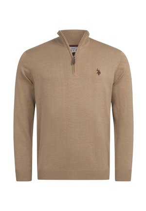 Mann lächelnd, trägt einen beigefarbenen Pullover mit Viertelreißverschluss und kleinem Logo auf der Brust und blaue Jeans, Hand in der Tasche.