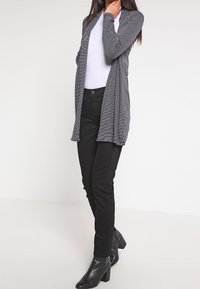 Šedý pruhovaný dlouhý kardigan přes bílou blůzu, v kombinaci s černými slim-fit kalhotami a černými botami na vysokém podpatku. Hladká textura, otevřený přední design.
