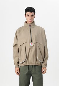 Beige vindjakke med høy krage, frontglidelås og reflekterende detaljer. Har store frontlommer og elastiske mansjetter.