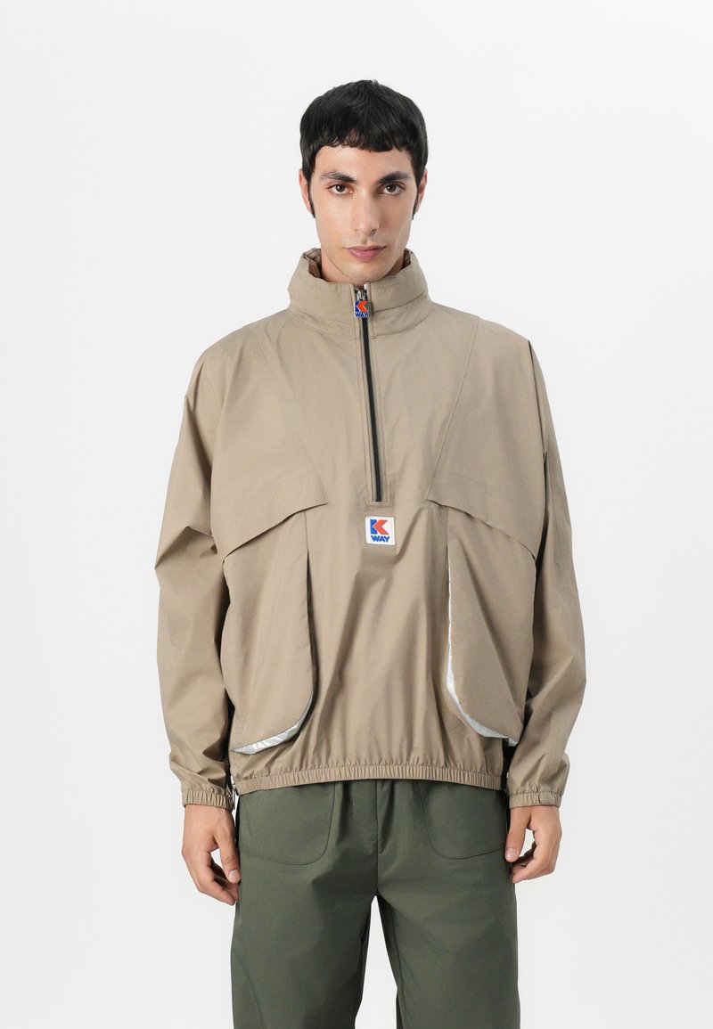 Beige windbreaker jacket with a high collar, front zipper, and reflective accents. Features large front pockets and elasticated cuffs.