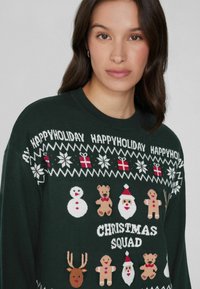 Grüner Strickpullover mit festlichen Grafiken: Schneemänner, Teddybären, Santa, Rentiere und dem Text "CHRISTMAS SQUAD" in Weiß.