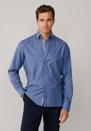 Camisa de manga larga abotonada en algodón azul claro con un sutil patrón texturizado, con botones marrones y un forro interior del cuello en contraste.
