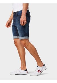 Tummansinisiä farkkushortsit, joissa on käännetty helma, yhdistettynä valkoisiin tennareihin, joissa on punaisia ja sinisiä yksityiskohtia. Iho näkyy paljailla jaloilla.