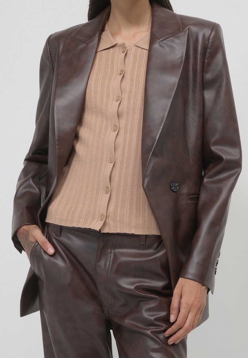Femme portant un blazer en cuir marron et un pantalon sur un top en maille côtelée beige à boutons, une main dans la poche et un tatouage visible sur le bras.