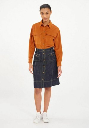 Chemise en velours côtelé marron avec deux poches poitrine, associée à une jupe en denim taille haute ornée de boutons et de coutures jaunes, portée avec des baskets blanches.