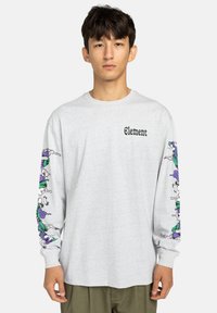 Junger Mann trägt ein hellgraues Langarmshirt mit dem Schriftzug "Element" und bunten abstrakten Designs an beiden Ärmeln, während er vor einem einfarbigen Hintergrund steht.
