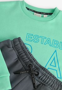 Grön sweatshirt med "ETABLERADE" tryckt i blått; grå joggers med texturerad midjeband och svart snörningsdetalj.