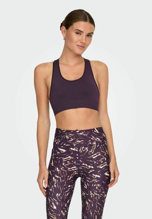 Woman with hair in a bun wearing a dark purple sports bra and high-waisted purple patterned leggings, standing against a plain background.