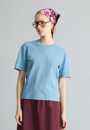 Kobieta w jasnoniebieskiej koszuli z krótkim rękawem, burgundowej spódnicy, dużych brązowych okularach i różowym kwiecistym chustce na głowie na szarym tle.