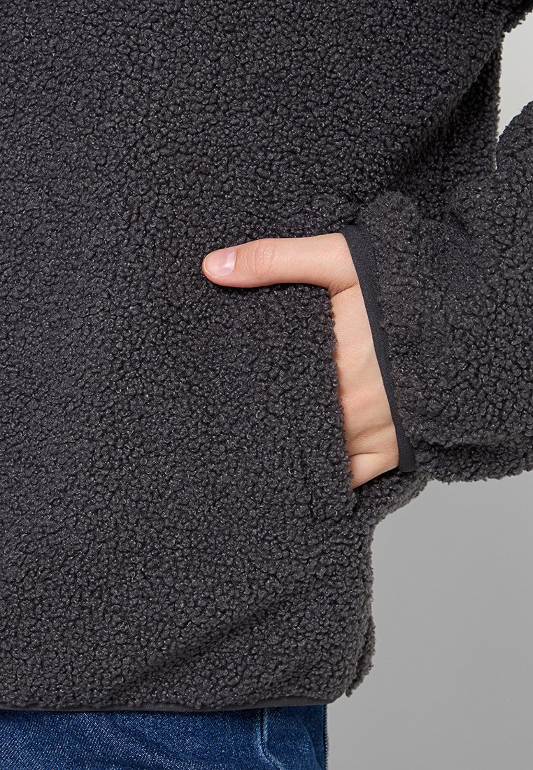 Dunkelgrauer Fleece-Pullover mit einer strukturierten, plüschigen Oberfläche. Verfügt über eine Seitentasche und enganliegende Bündchen. Sichtbare Hand, die in die Tasche eingeführt ist.