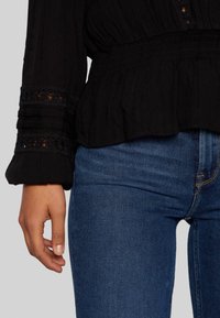 Mustassa tekstuurilla varustettu pusero, jossa on kerrostettu vyötärö ja pitsireunus, yhdistettynä sinisiin farkkuihin. Paino on hihnoissa, joissa on röyhelöyksityiskohtia, rento malli.