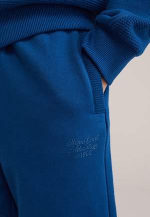 Blå joggingbukser lavet af blødt stof med ribbede manchetter, teksturerede detaljer og et diskret logo præget på frontlommen.