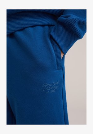 Pantalones de chándal azules hechos de tela suave, con puños acanalados, detalles texturizados y un logo sutil en relieve en el bolsillo delantero.