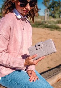 Žena s brýlemi, růžovým svetrem a džínami, která drží světle šedou prošívanou peněženku s klipsou, sedí venku na dřevěné lavičce.