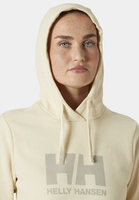 Sudadera color crema con un bolsillo frontal, cordones y un gran logo gris "HH". Tejido suave con una textura lisa, diseño casual.