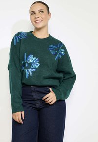 Pullover verde a maglia con design floreali in paillettes blu sulle spalle, abbinato a jeans scuri a vita alta.